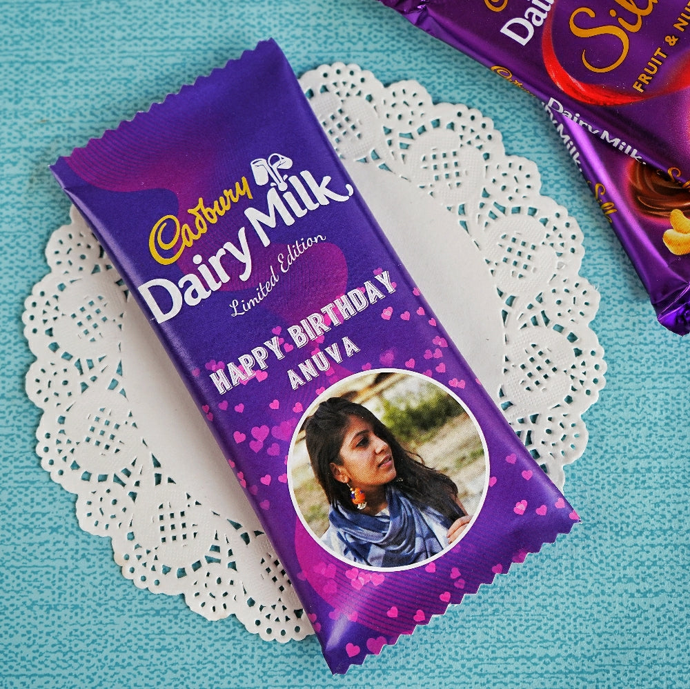 A Dairy Milk chocolate bar wrapped in customized packaging with a photo and text on it, placed on a white doily.