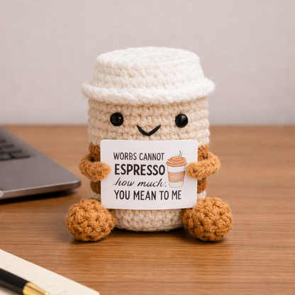 Crocheted coffee cup with a face and humorous message on a wooden surface.