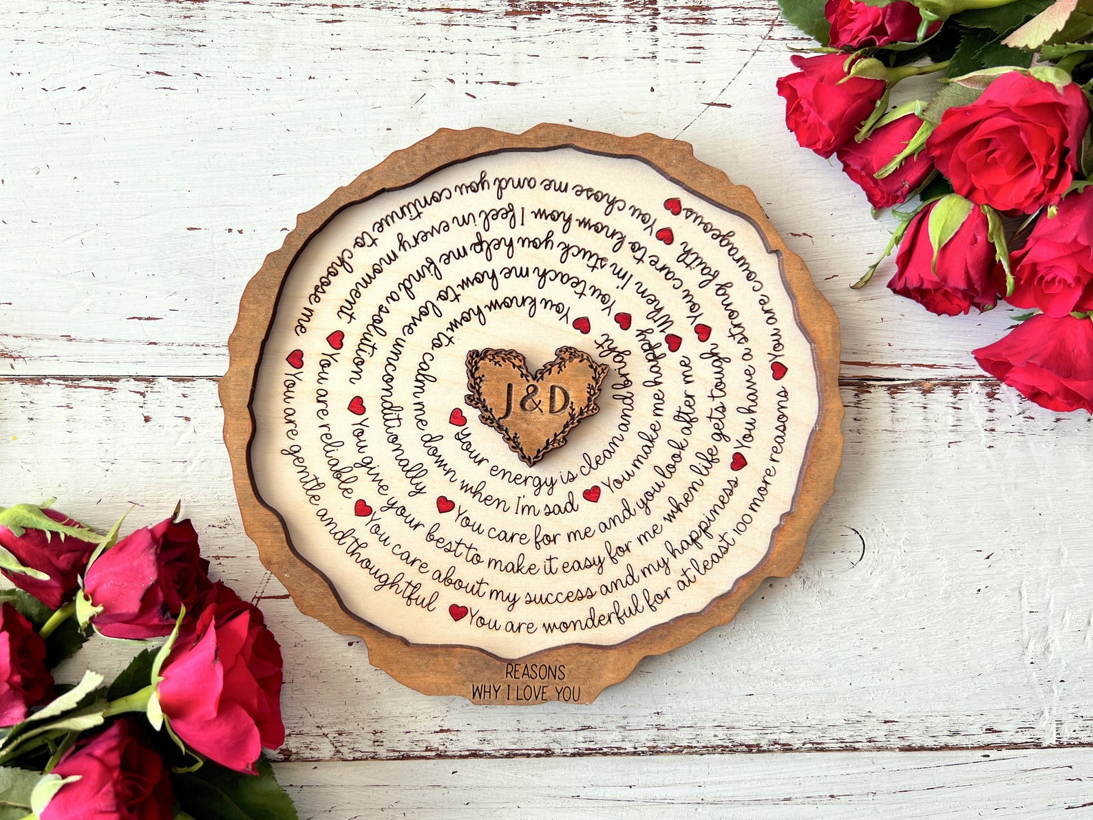 Wooden plaque with engraved text and a heart-shaped cutout, surrounded by red roses on a rustic wooden surface.